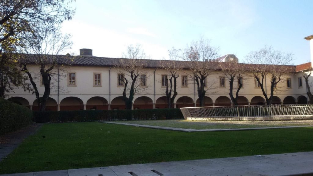 A long, historic building borders a quiet courtyard with a lawn, leafless trees, and a circular fountain