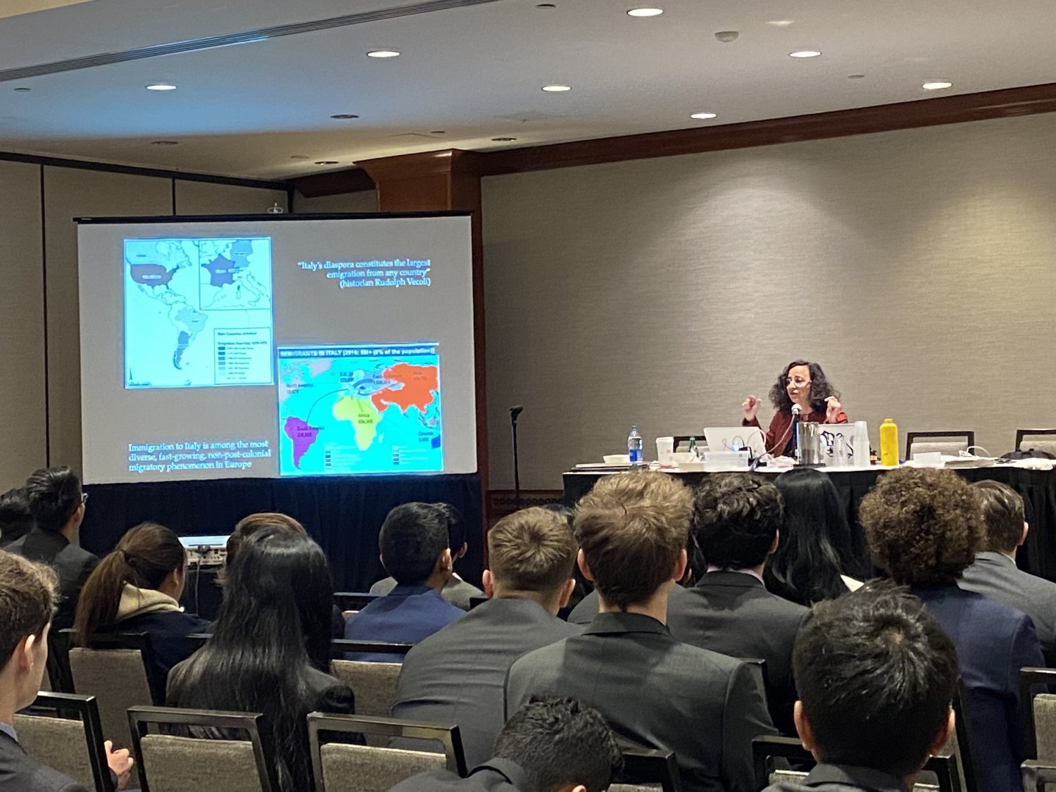 A woman sits behind a table in a ballroom and speaks to a crowd of college students about an image on a projector screen