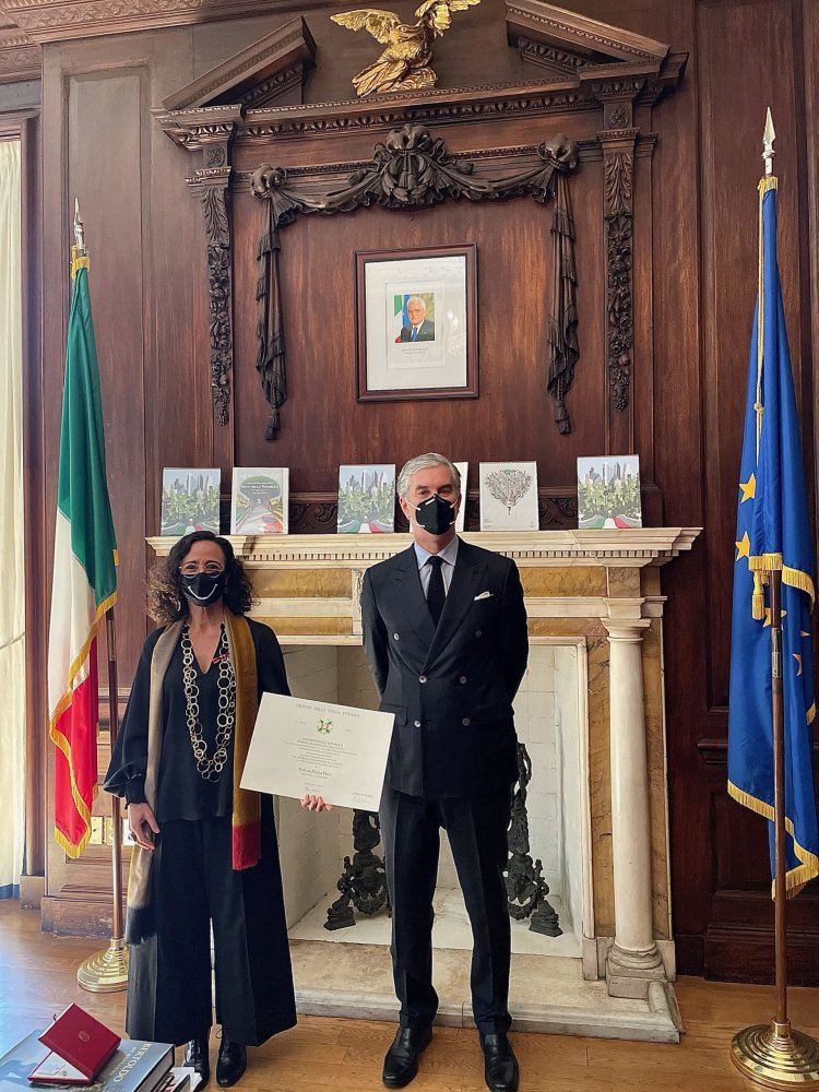 Two people stand in front an ornate mantel between an Italian flag and an EU flag and one person holds a paper certificate