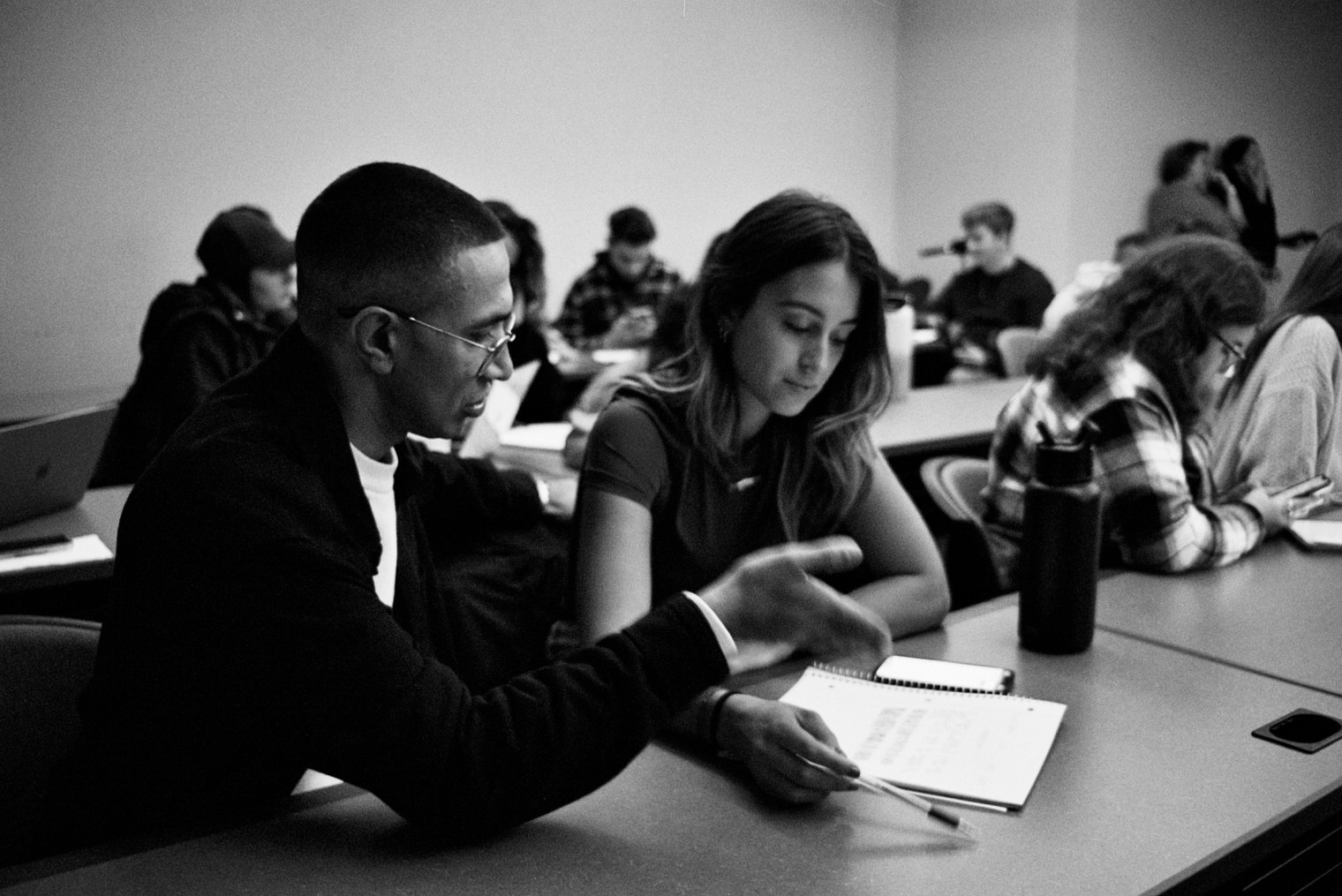 A black and white image of a man sitting beside a student in a classroom discussing an assignment