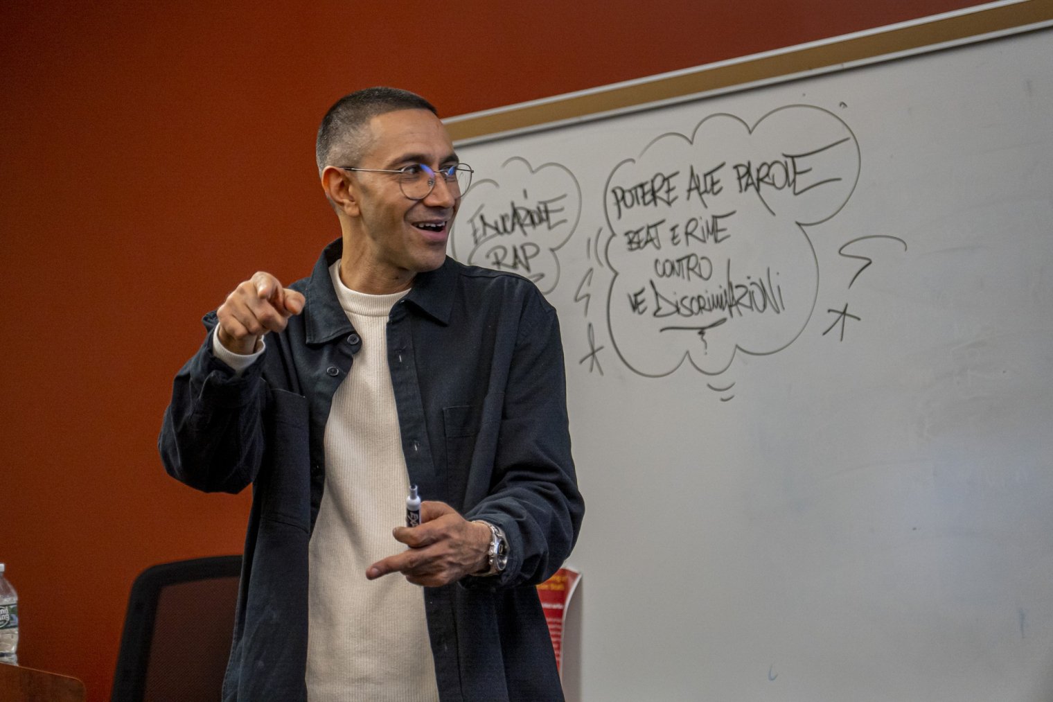 A man holds a marker and stands in front of a whiteboard with writing on it  