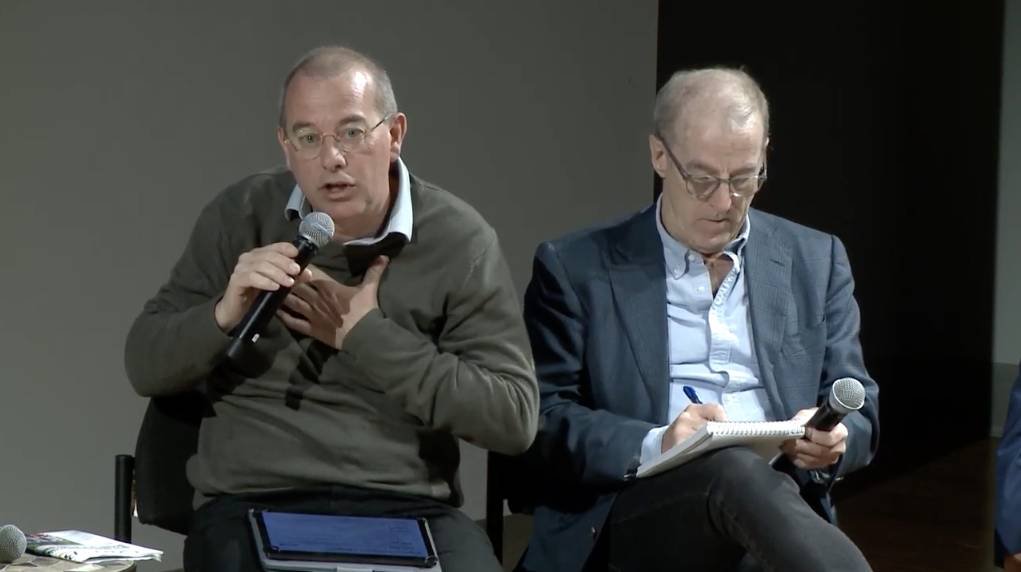 Two people sit on a stage, one hold a microphone and speaks while another writes notes into a notebook