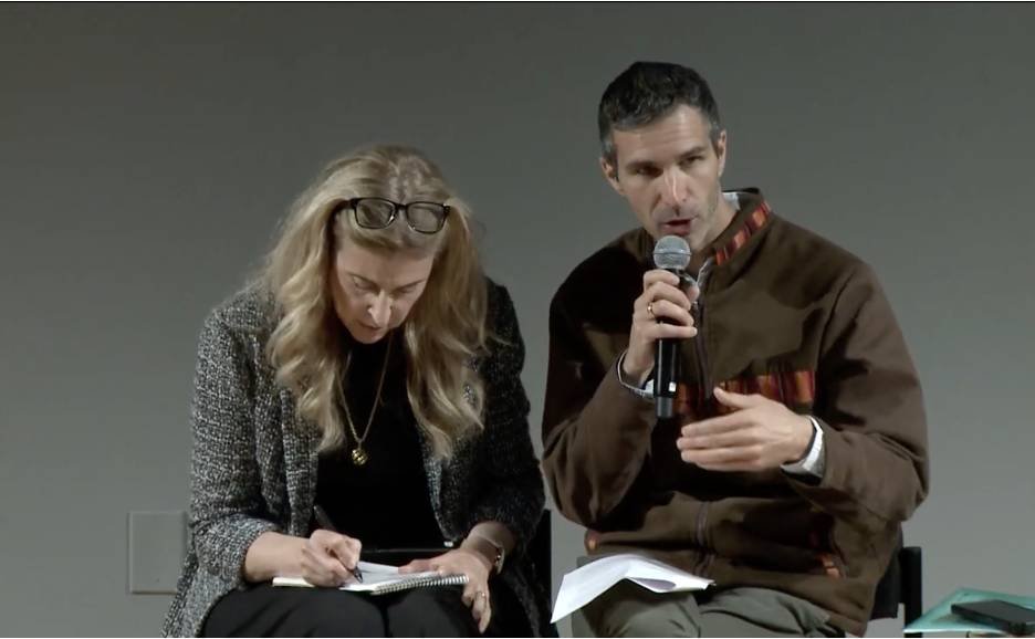 Two people sit on a stage, one hold a microphone and speaks while another writes notes into a notebook