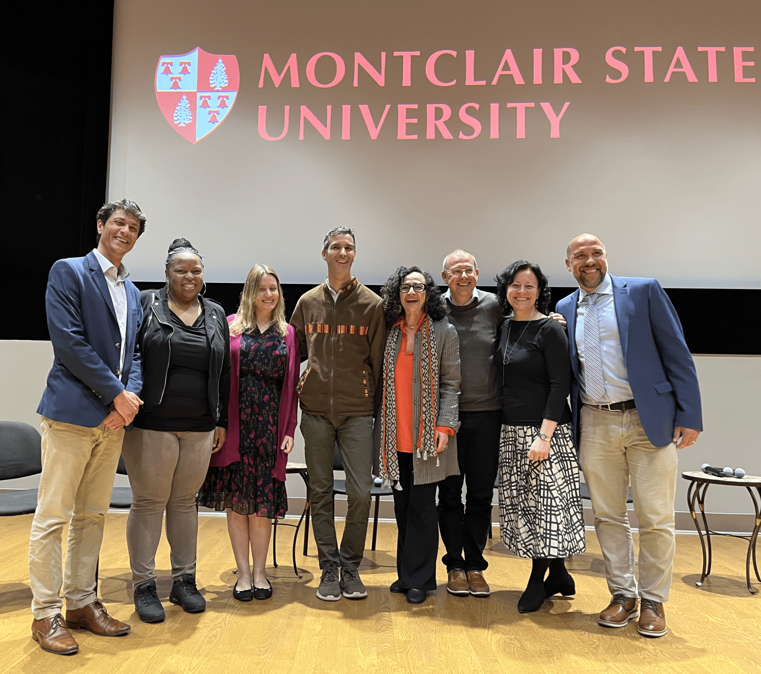 A group of people pose together in front of a projection screen bearing the words "Montclair State University"