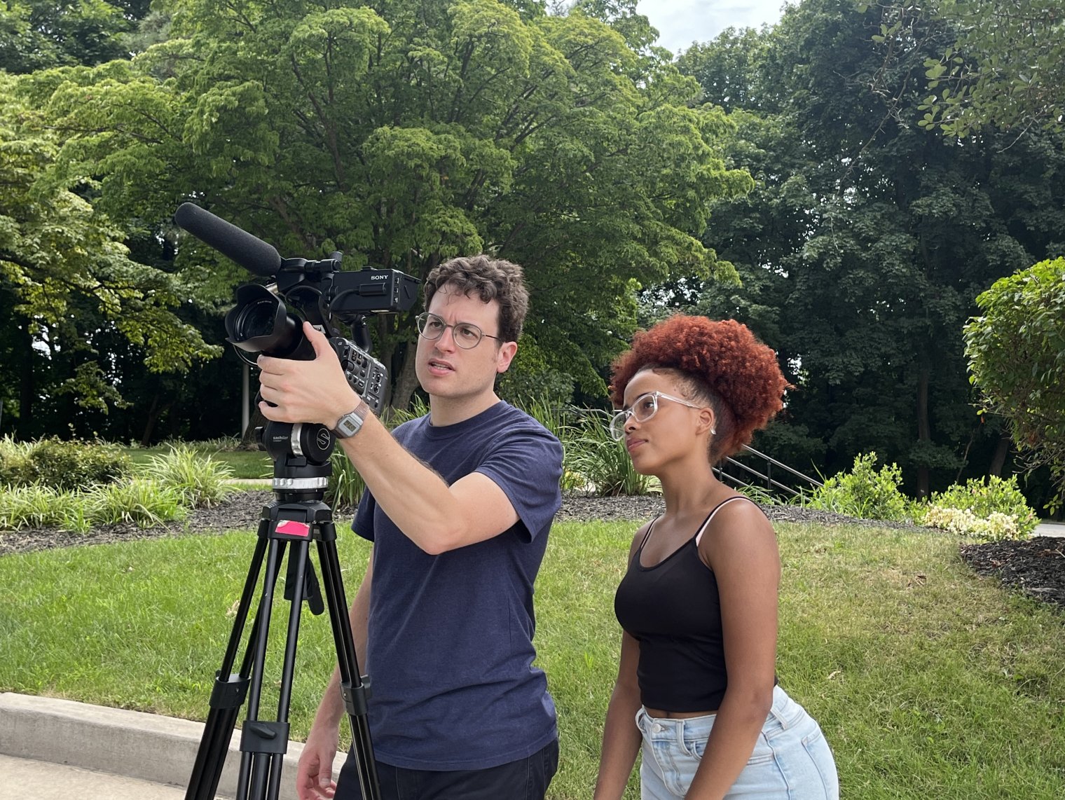 One person demonstrates how to use a video camera on a tripod to another person