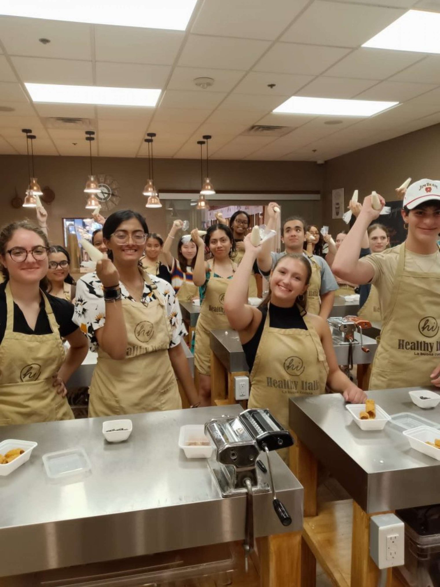 A group of college students wear matching aprons and pose in a kitchen holding pastry basters