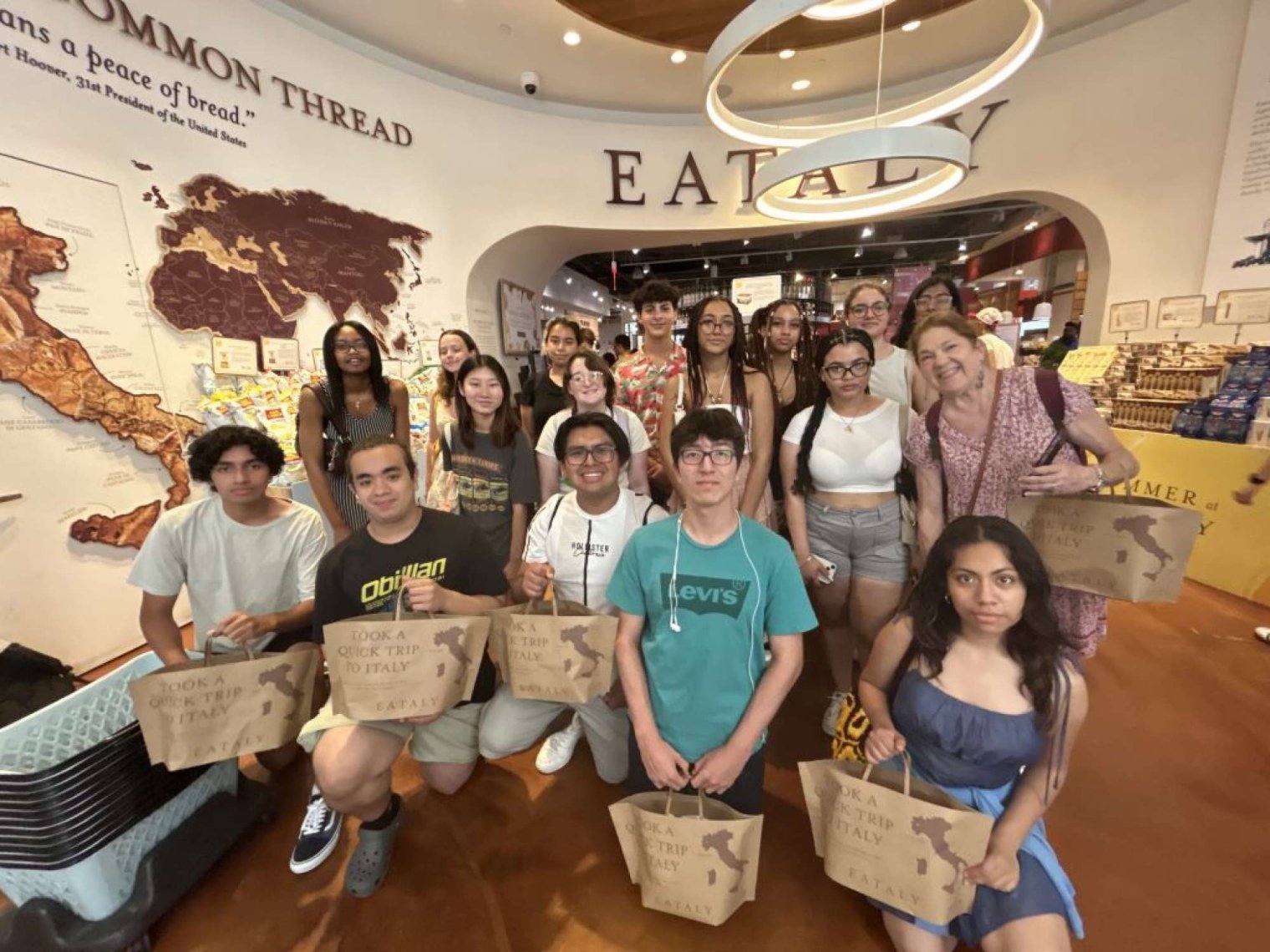 A group of college students pose with bags in front a wall with a world map on it and the name "Eataly"