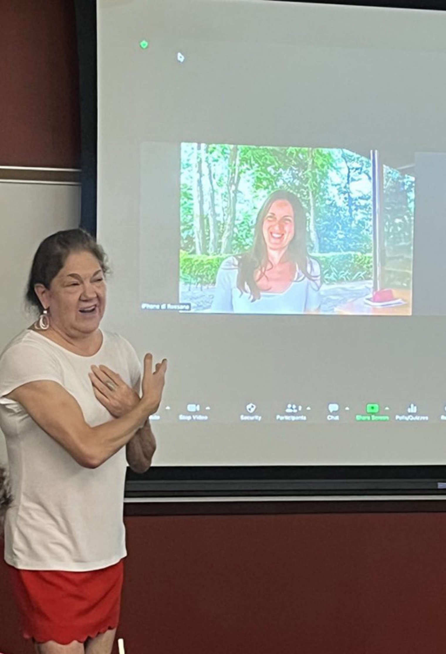 A woman stands beside a projector screen and points to the image on the screen and addresses an audience