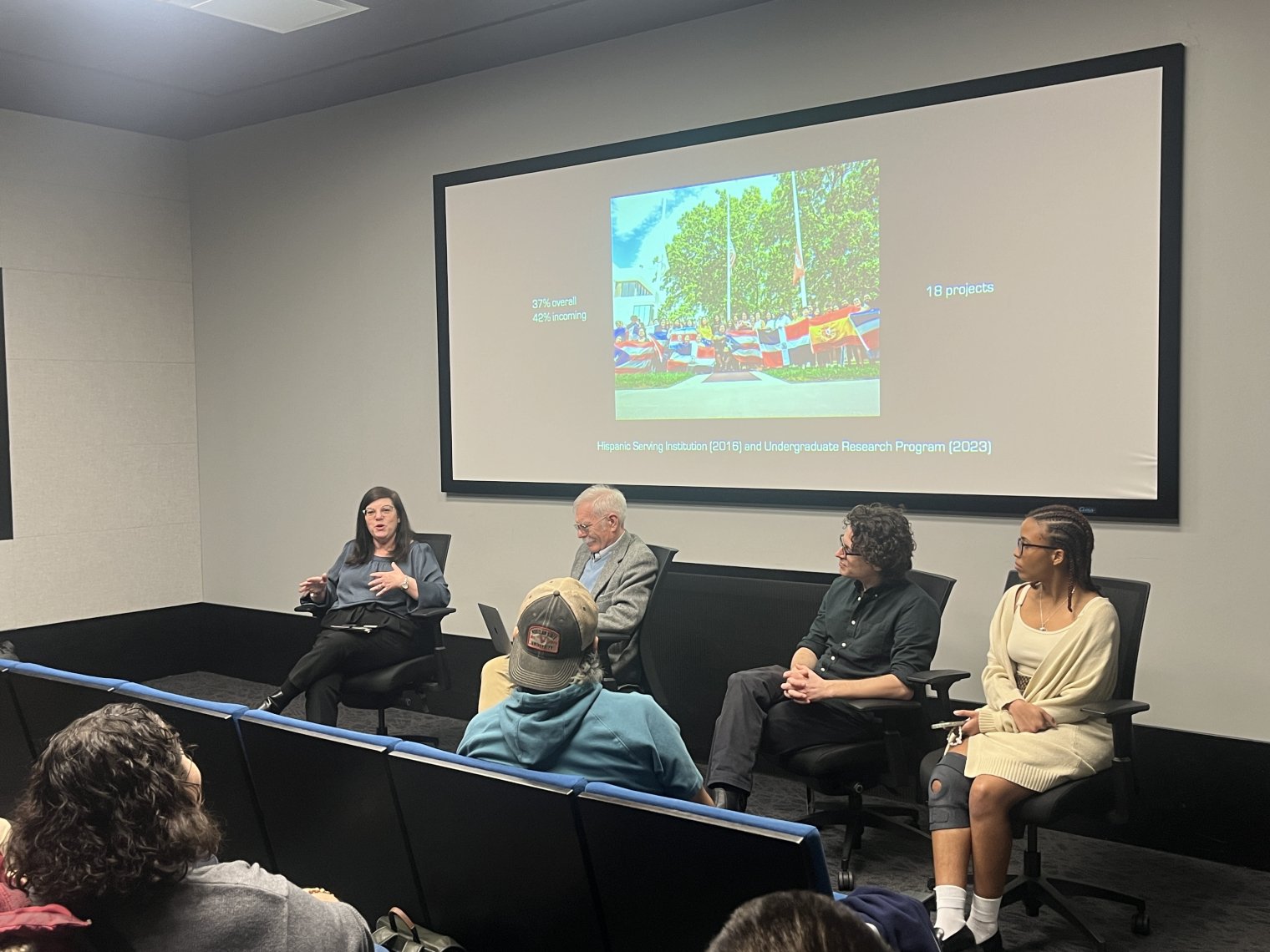 A panel of four people present to an audience in front of a projector screen