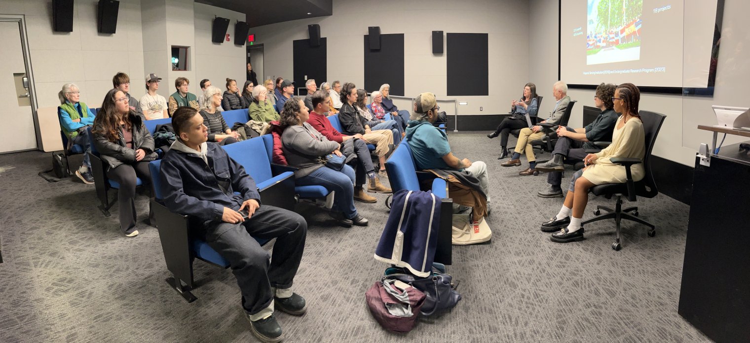 An audience sits in theatre seats and listen to a presentation by four panelists