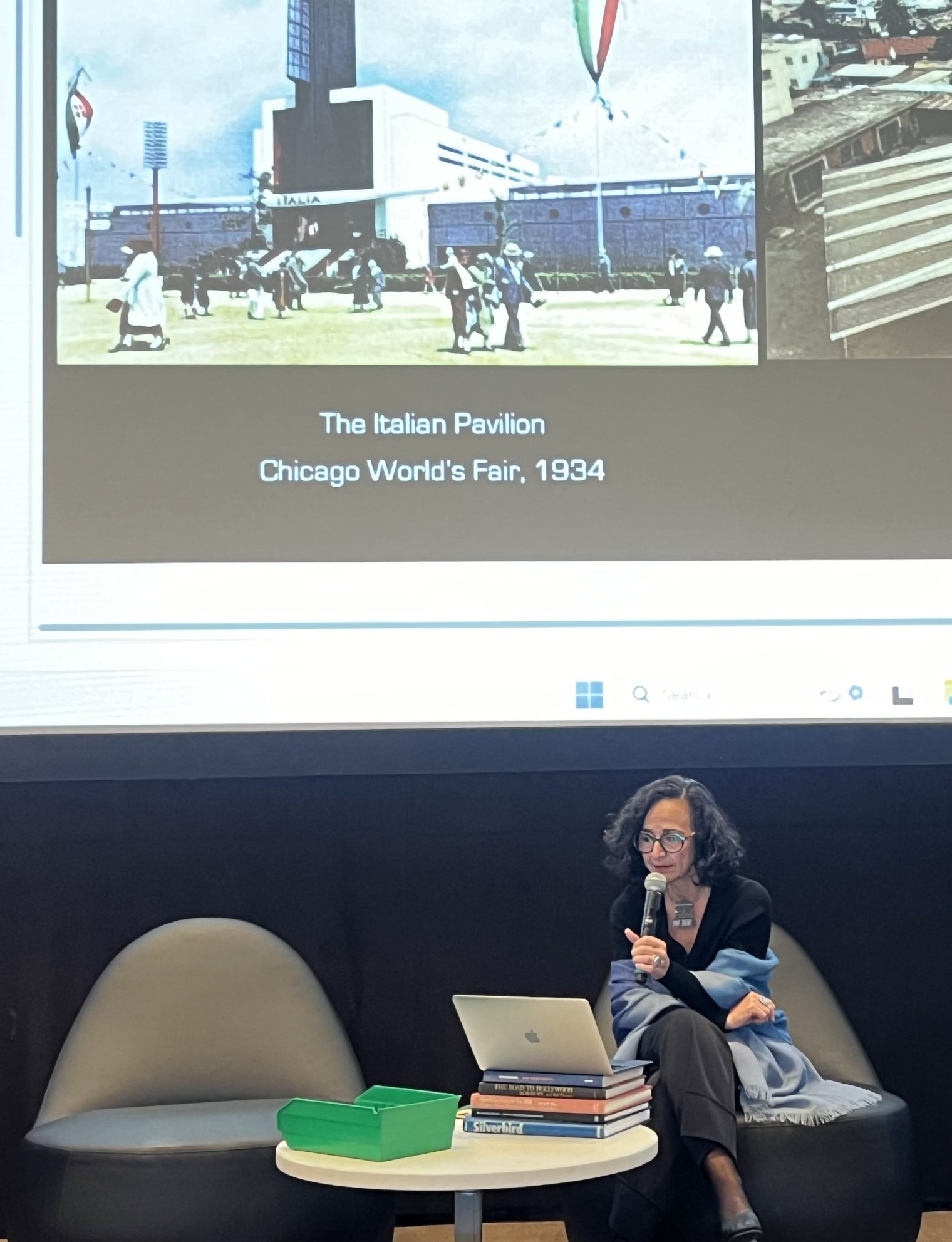 A woman sits at a laptop and delivers a presentation in front of a projector screen