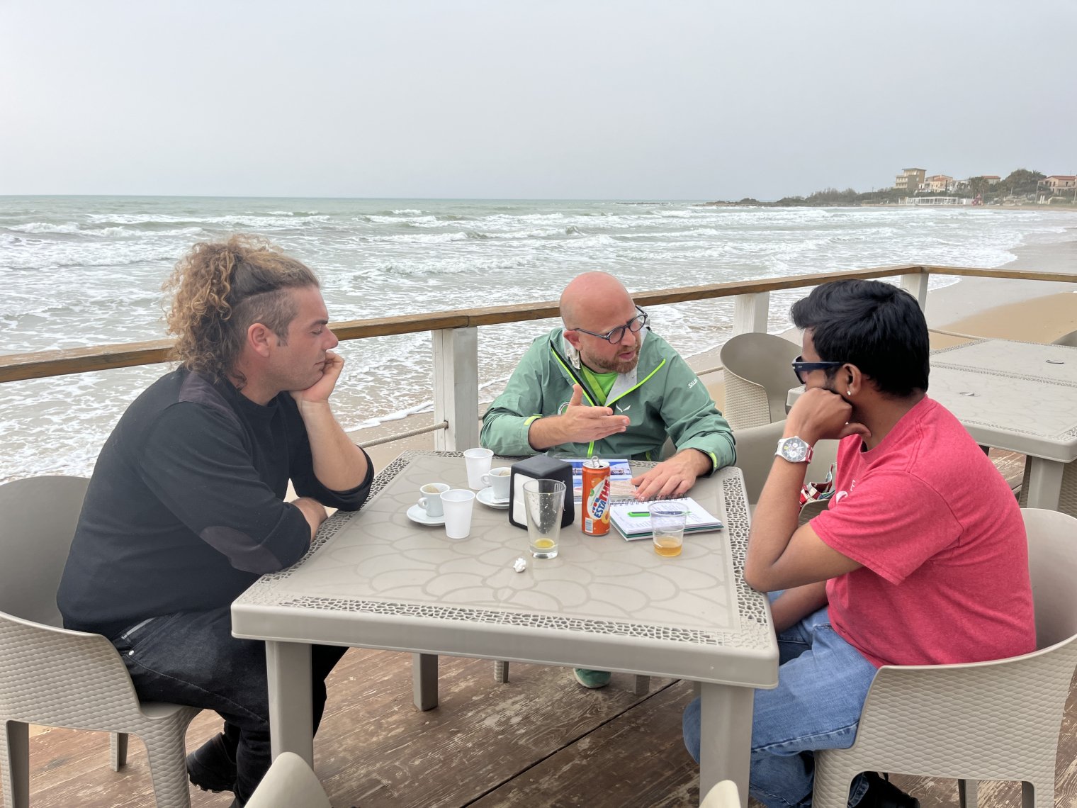 Three people sit around a table by the ocean and talk