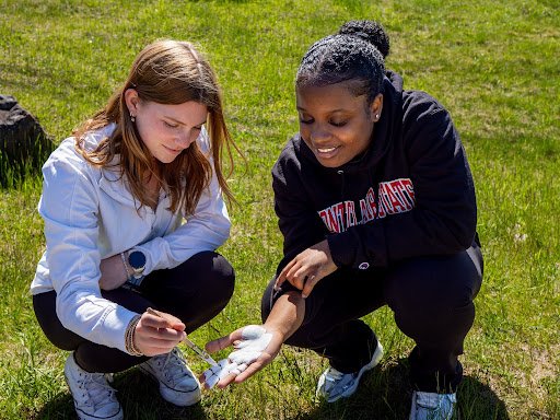 Two students on grass while one is painting the others hand with white paint.