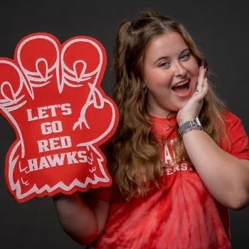 Woman student in red shirt holding a foam talon that reads Let's Go Red Hawks