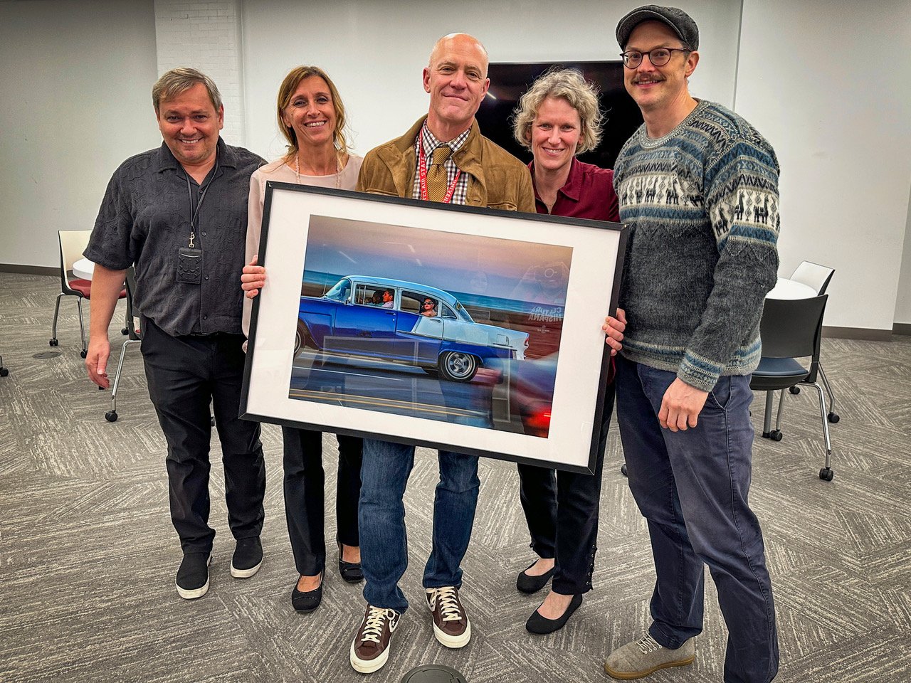 A group of people stand and pose in a conference room, the person in the middle holds a large, framed photograph