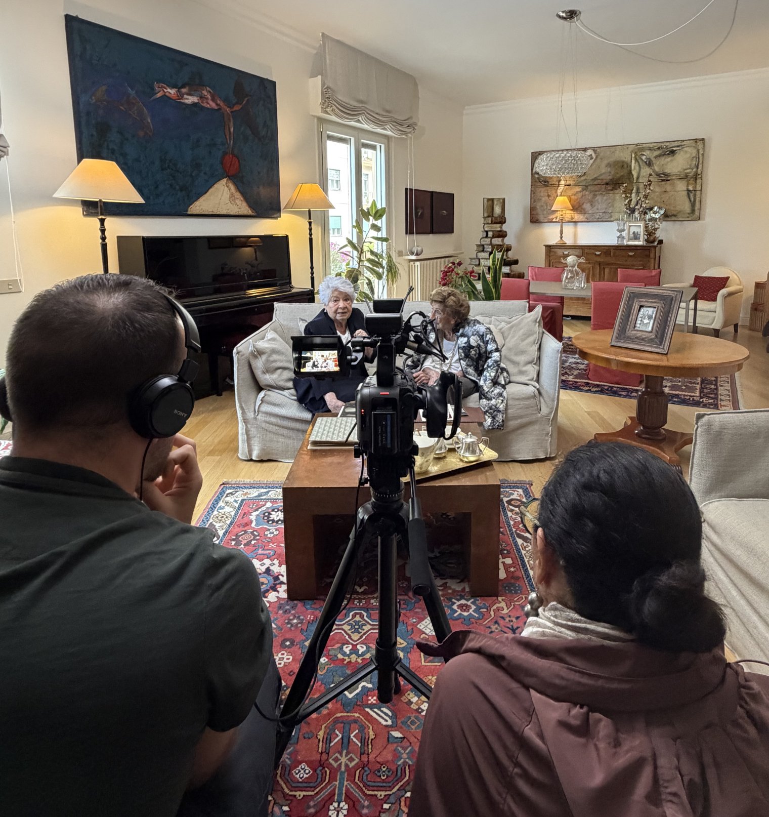 Two people sit behind a camera filming two people sitting on a couch inside a house