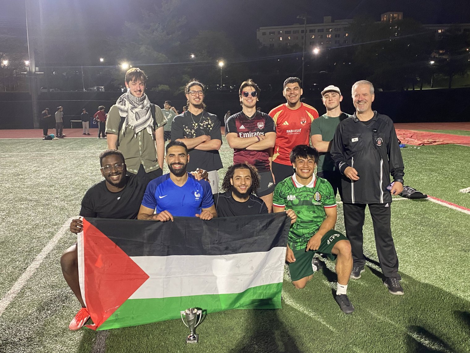 A group of college students pose on a soccer pitch on night, several hold up a large Palestinian flag