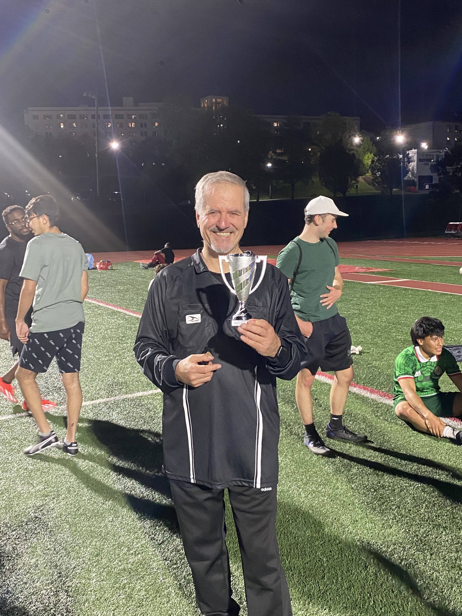 A man stands on a soccer pitch at night holding a small golden trophy