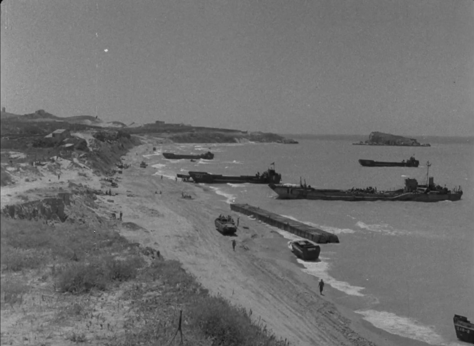 Black-and-white photo of wartime landing craft and ships along a sandy beach.