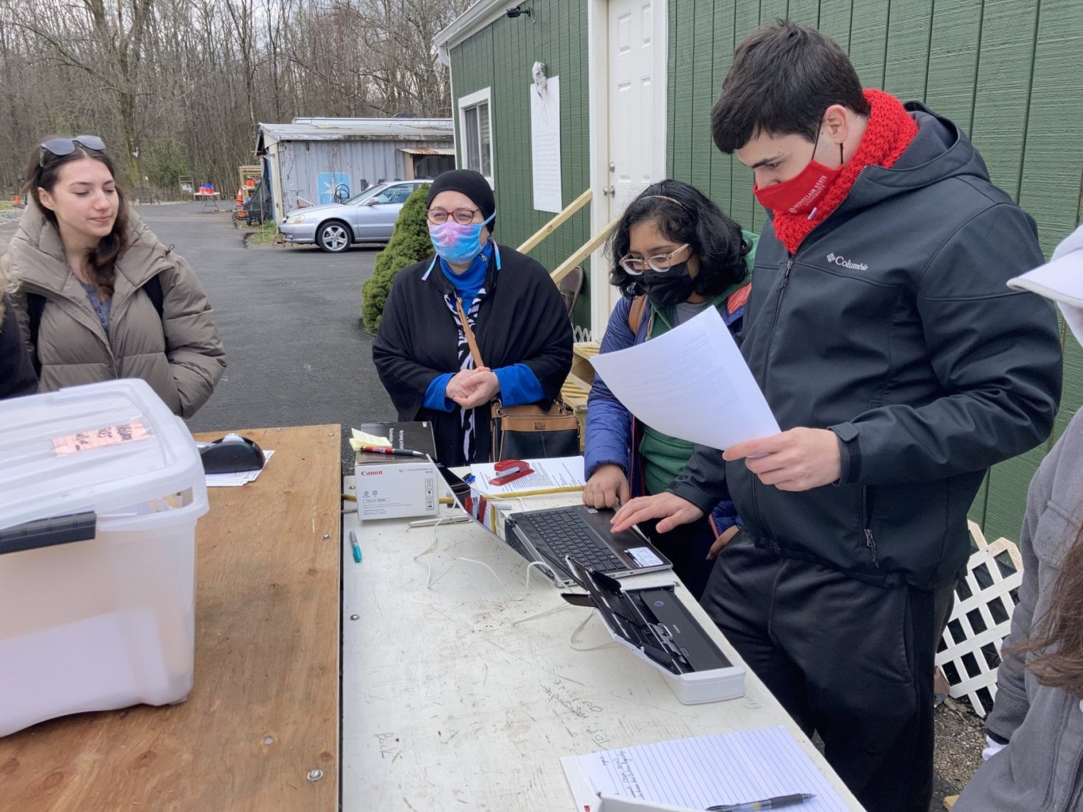 Several people wearing face masks stand around a table with a laptop and a printer
