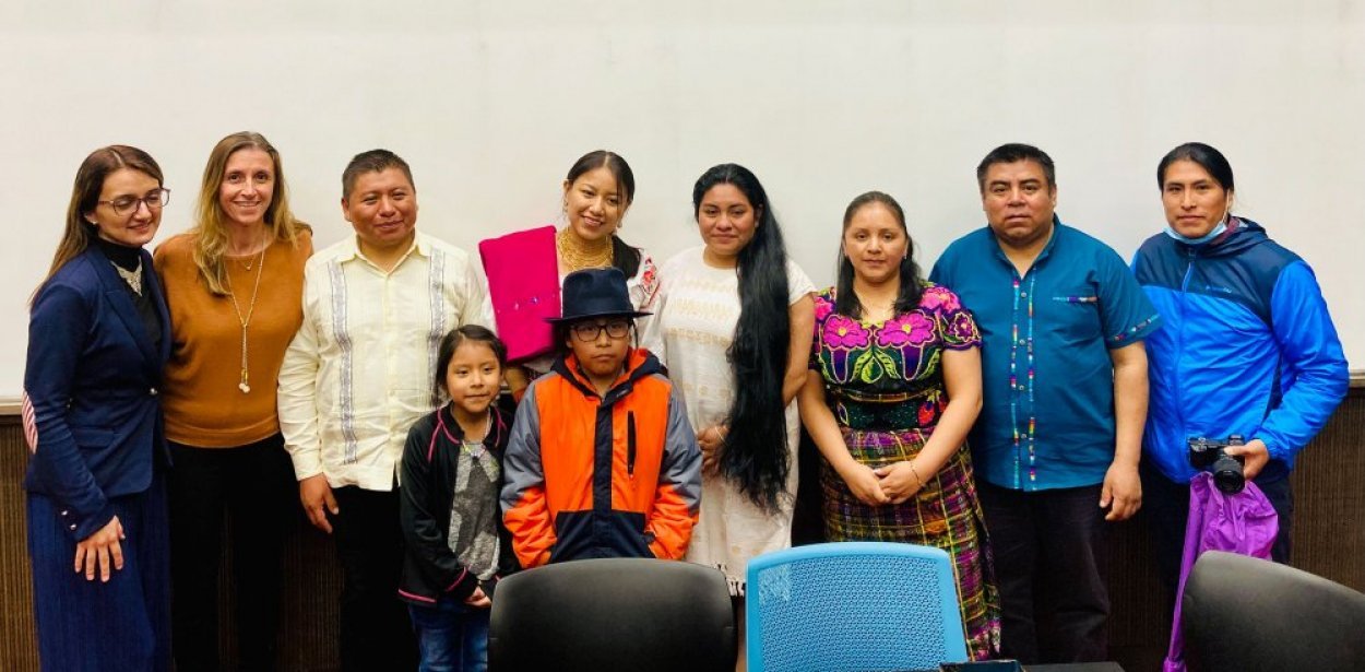 Group photo of adults and two children posing indoors. Several people wear colorful traditional or embroidered clothing, while others wear casual or business attire; one person holds a camera