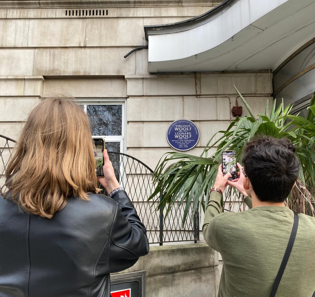 Two people take photos on their cell phones of a round plaque hanging on a stone wall