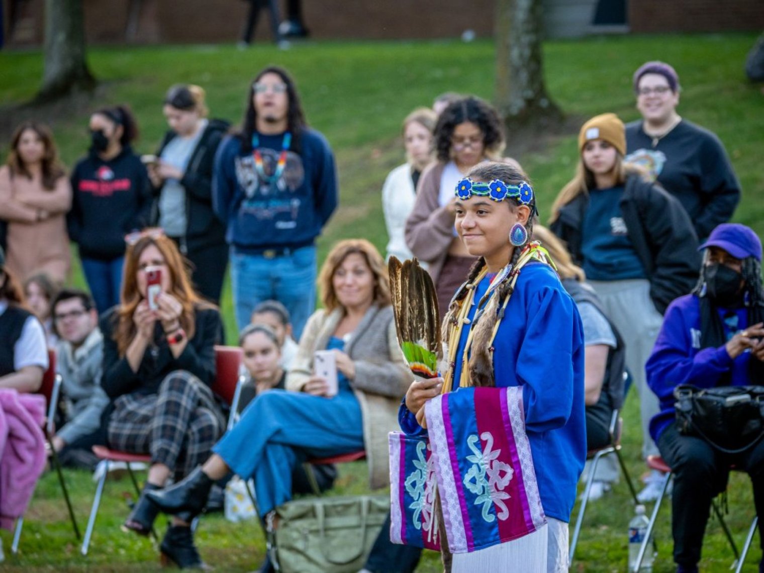 A young person in blue ceremonial regalia holds feather fans before a seated outdoor audience