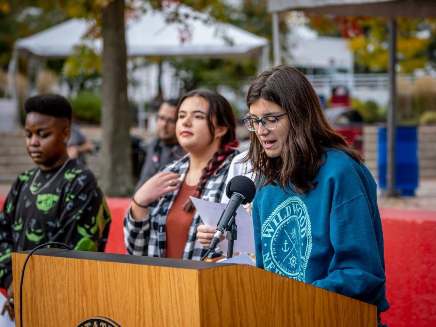A student reads from notes at an outdoor podium as others stand behind them