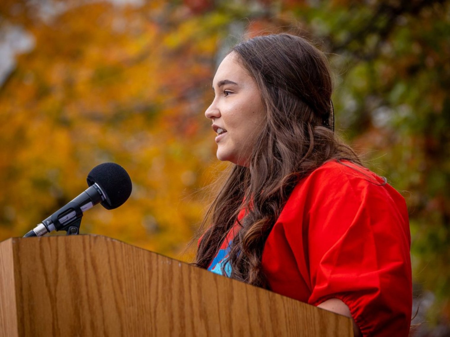 A woman in red speaks at an outdoor podium against a backdrop of autumn foliage