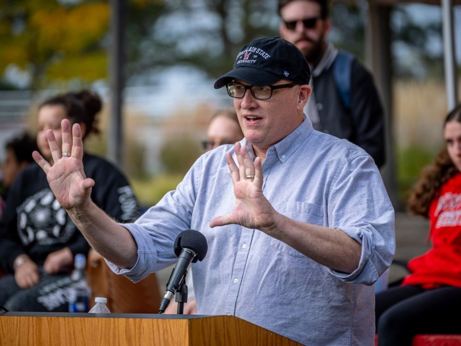 A man in a blue shirt and a ball cap speaks at an outdoor podium