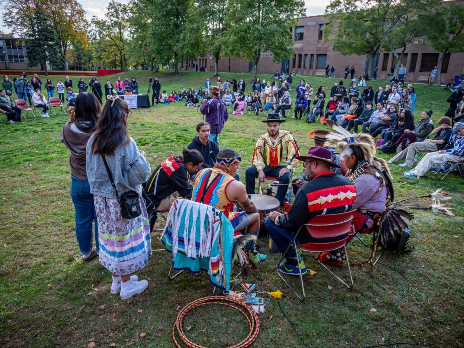 Indigenous performers gather around a drum before an outdoor campus audience
