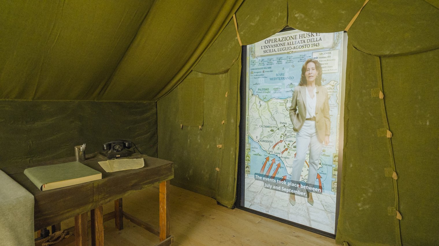 Interior of a WWII-style military tent exhibit with a desk, telephone, documents, and a video display about Operation Husky