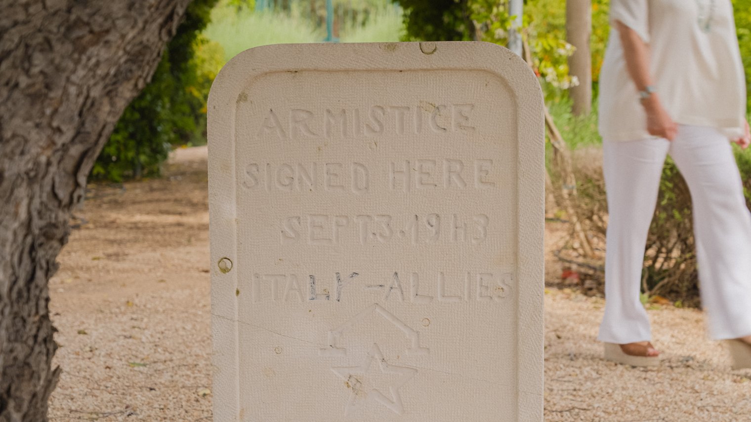 Close-up of a stone marker reading “Armistice signed here Sept. 3, 1943, Italy–Allies,” with a person walking behind it