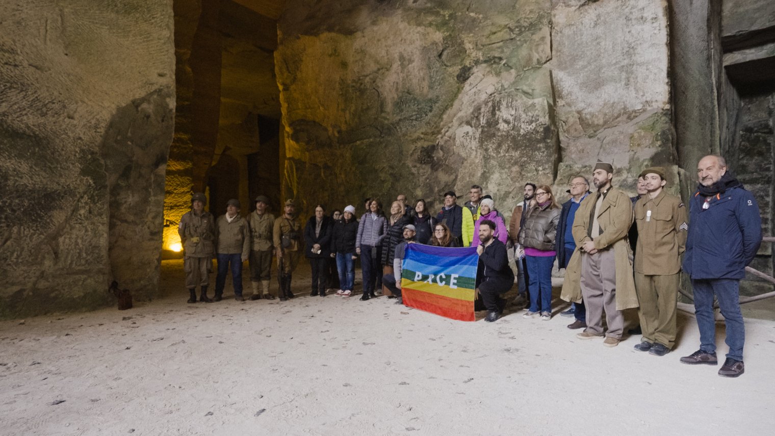 A large group of people stand in a line, holding a rainbow flag with the words "Peace"