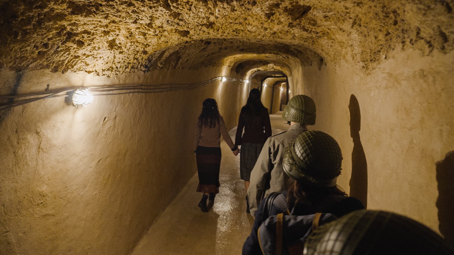 A group of people, including men in military attire, walk through a cave lit by lightbulbs