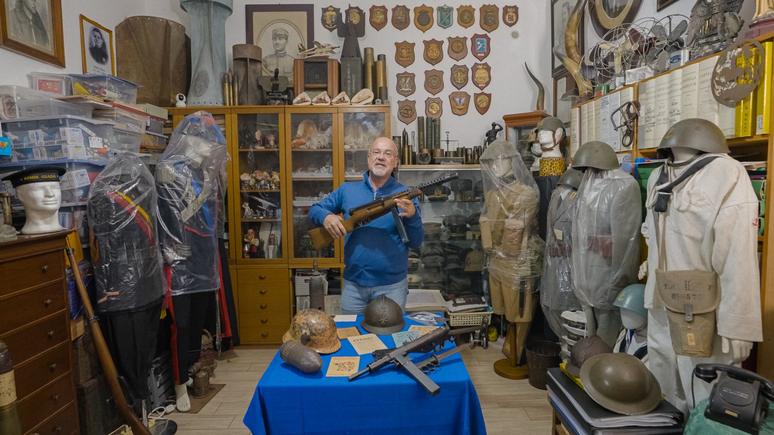A man stands in a room full of vintage military artifacts, holding an antique firearm