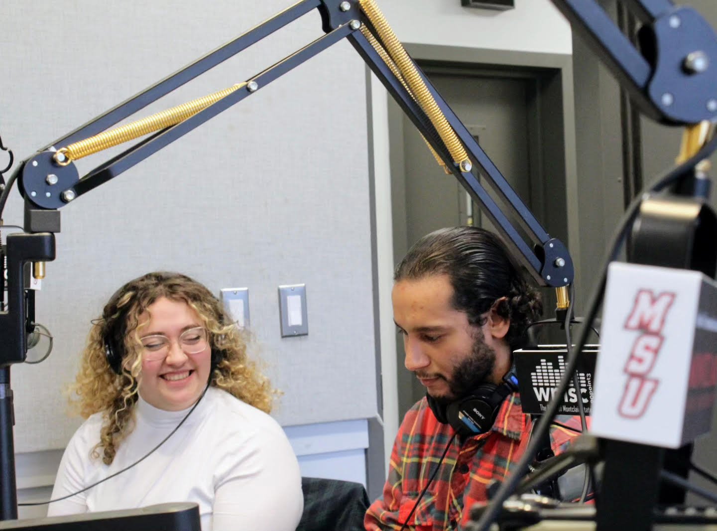 Two people sit in a radio booth near a microphonr, one wears headphones