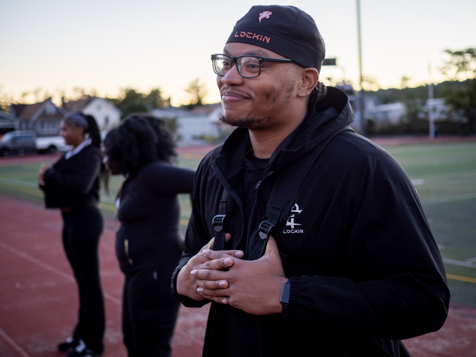  Montclair alumnus Emmanuel Ozias ’25 smiles while standing on the track beside a football field, wearing a black Lock1n jacket and cap. 