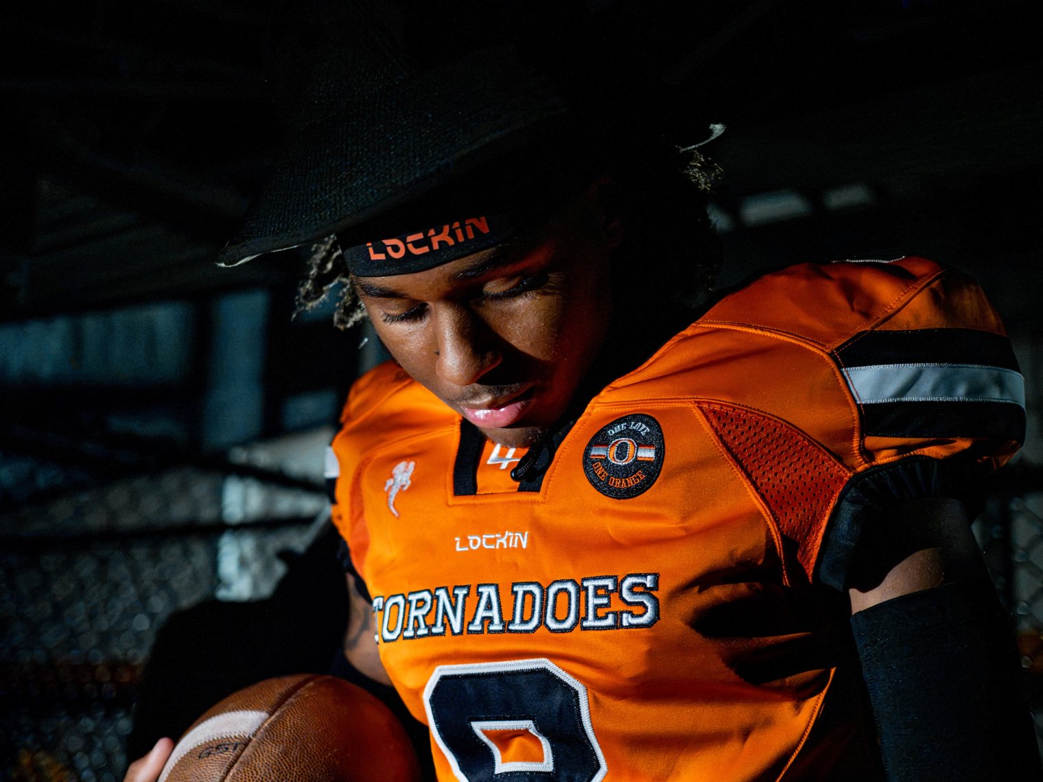 An Orange High School football player looks down at a football in his hand while wearing a bright orange Lock1n jersey with “Tornadoes” across the chest.