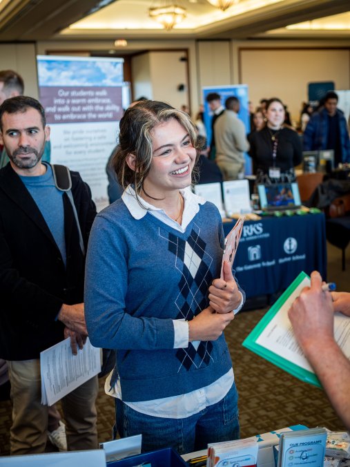 a woman smiles while talking to prospective employers at a career fair, holding papers in her hand