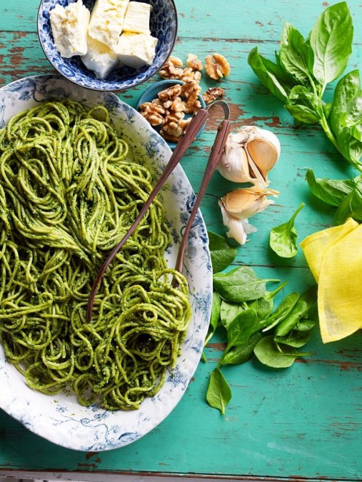 a counter has a large bowl of pesto linguine with serving utensil. ingredients used to make the pesto (cheese, fresh basil, nuts, garlic) are on the counter next to the bowl