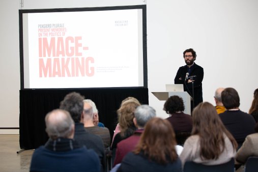 A person stands at a podium, beside a projection screen and speaks to a crowd