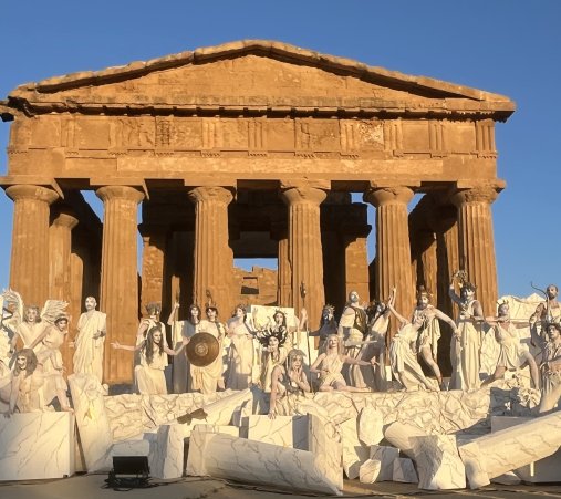 A group of actors in classical-era Greek togas pose in front of a temple