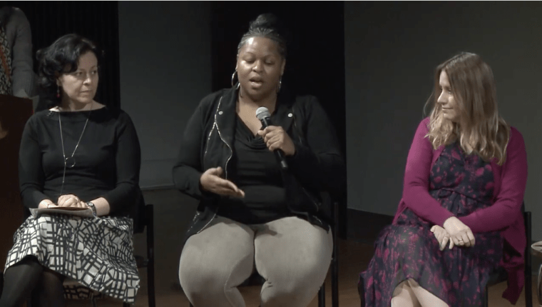 Three people sit in chairs on a stage, one is holding a microphone and speaking while the other two watch