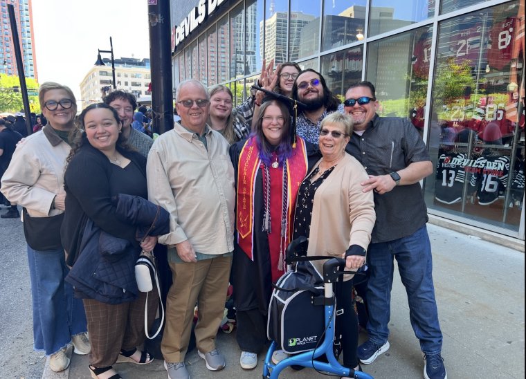 A group of people of varying ages pose together outside around a woman wearing a black cap and gown