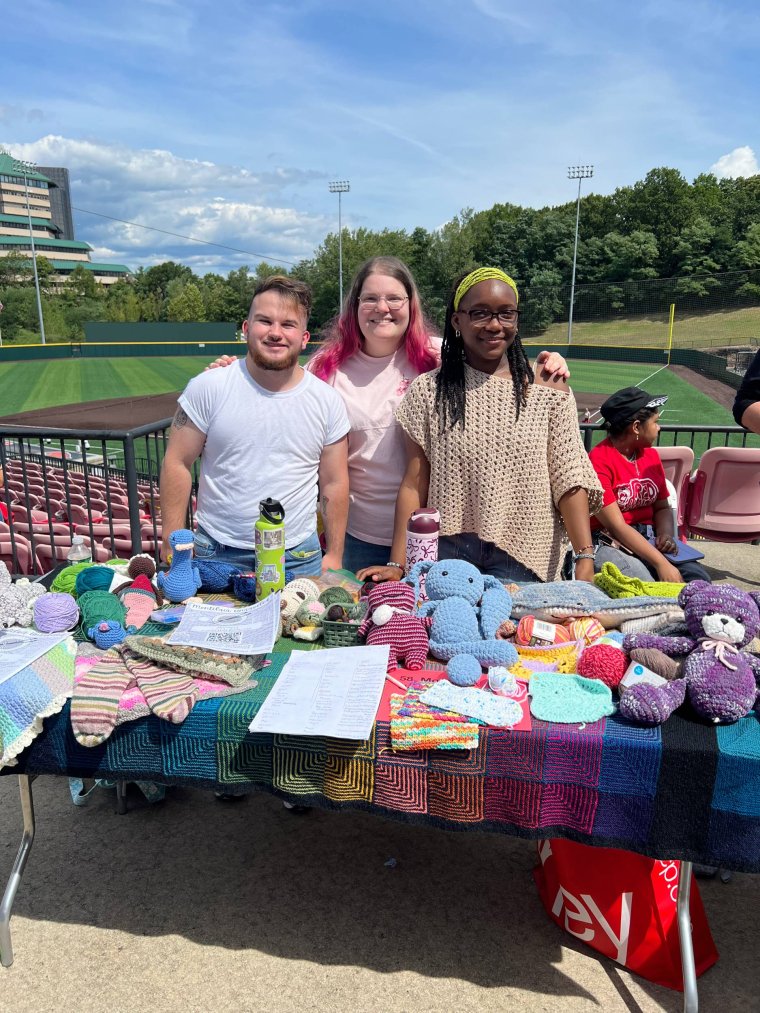 Three college students pose at a baseball park standing behind a table of knitted items 